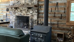 A rustic cabin interior with a stone fireplace, a black wood stove labeled 'FISHER', wooden walls, and a leather chair. - Retreat with rustic cabin, picnic shed and ponds (Leicester)
