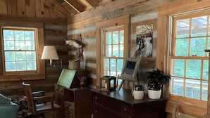 A rustic cabin interior with wooden walls, a desk with a computer, a chair, a lamp, and a window with a view of trees. - Retreat with rustic cabin, picnic shed and ponds (Leicester)