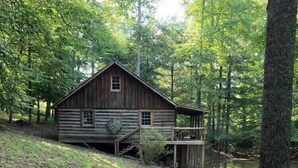 A rustic cabin with a wooden deck, surrounded by dense forest and a gravel path leading to it. - Retreat with rustic cabin, picnic shed and ponds (Leicester)