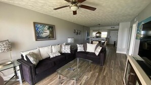 A living room with a sectional sofa, a glass coffee table, a ceiling fan, and a painting on the wall.