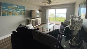 A living room with a sofa, coffee table, and a television on a cabinet. There is a balcony with a view of the water.