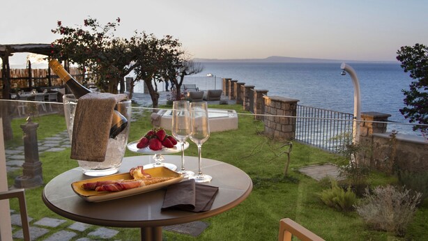 Eine Terrasse mit Tisch für zwei Personen, komplett mit Champagnerflasche, zwei Gläsern, einer Erdbeerenplatte und einem Hotdog. Die Terrasse blickt auf ein Gewässer und ist von Steinmauern und Grünpflanzen umgeben.