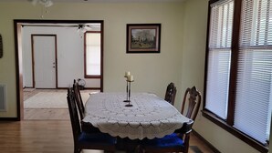 Uma área de refeições com uma mesa posta para quatro pessoas, um ventilador de teto, uma porta que dá para outro quarto e uma janela com persianas.