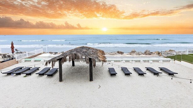 A beachside view with a thatched structure, sun loungers, and a white fence.