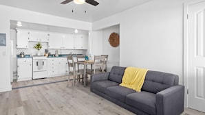 A modern kitchen with white cabinets, a dining area with a wooden table and chairs, and a gray sofa with a yellow throw blanket.
