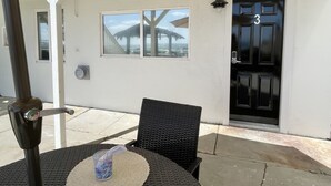 A covered patio area with a table and chairs, a black door with the number 3, and a window overlooking a beach. - Lower Level Pet Friendly Oceanfront Studio P925-3 (Oceanside)