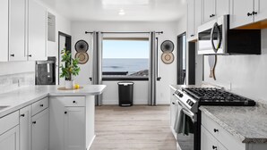 Una cucina moderna con mobili bianchi, un'isola centrale, un fornello e vista sull'oceano attraverso una grande finestra.