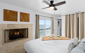 A Seashell Bedroom with a fireplace, a ceiling fan, a bed with a view of the ocean, and a balcony with curtains.