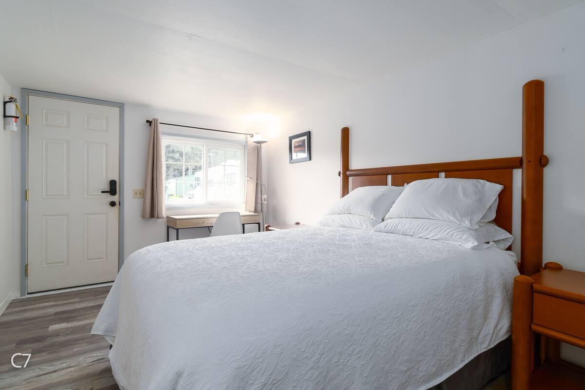 A bedroom with a bed, a wooden nightstand, a desk with a chair, a window with curtains, and a door.
