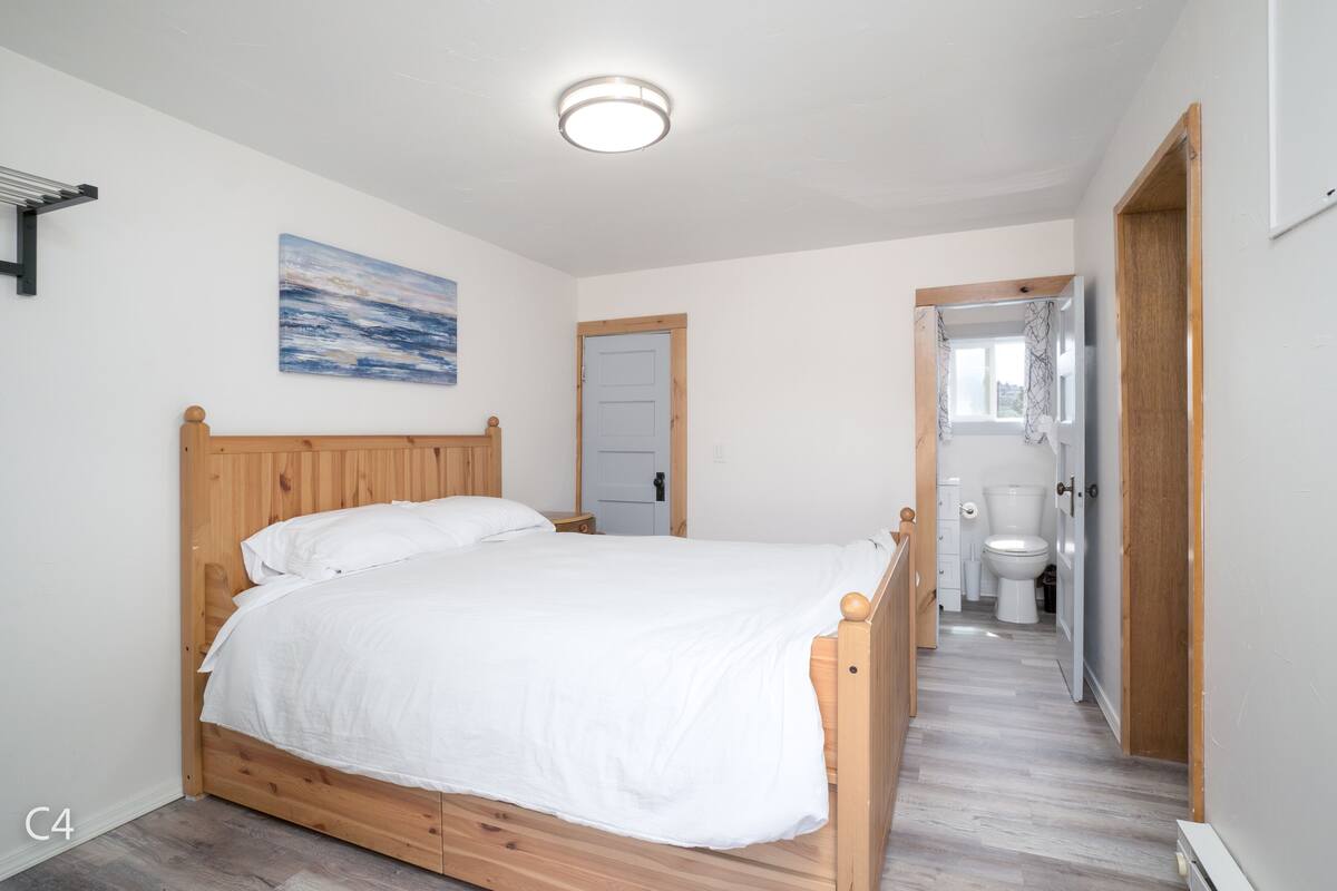 A bedroom with a wooden bed, a white wall, a painting, a door, and a bathroom visible through an open door.