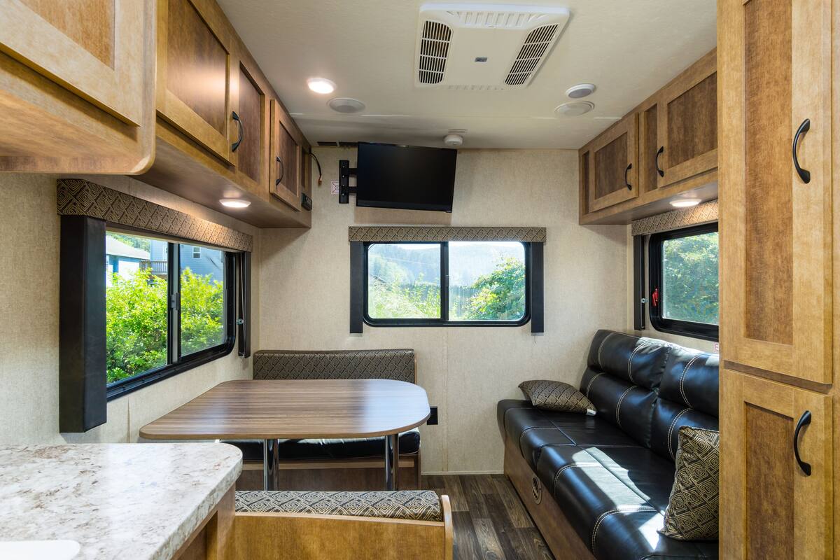 A compact RV interior with a dining area, a seating area with a sofa, and a window providing a view of greenery.