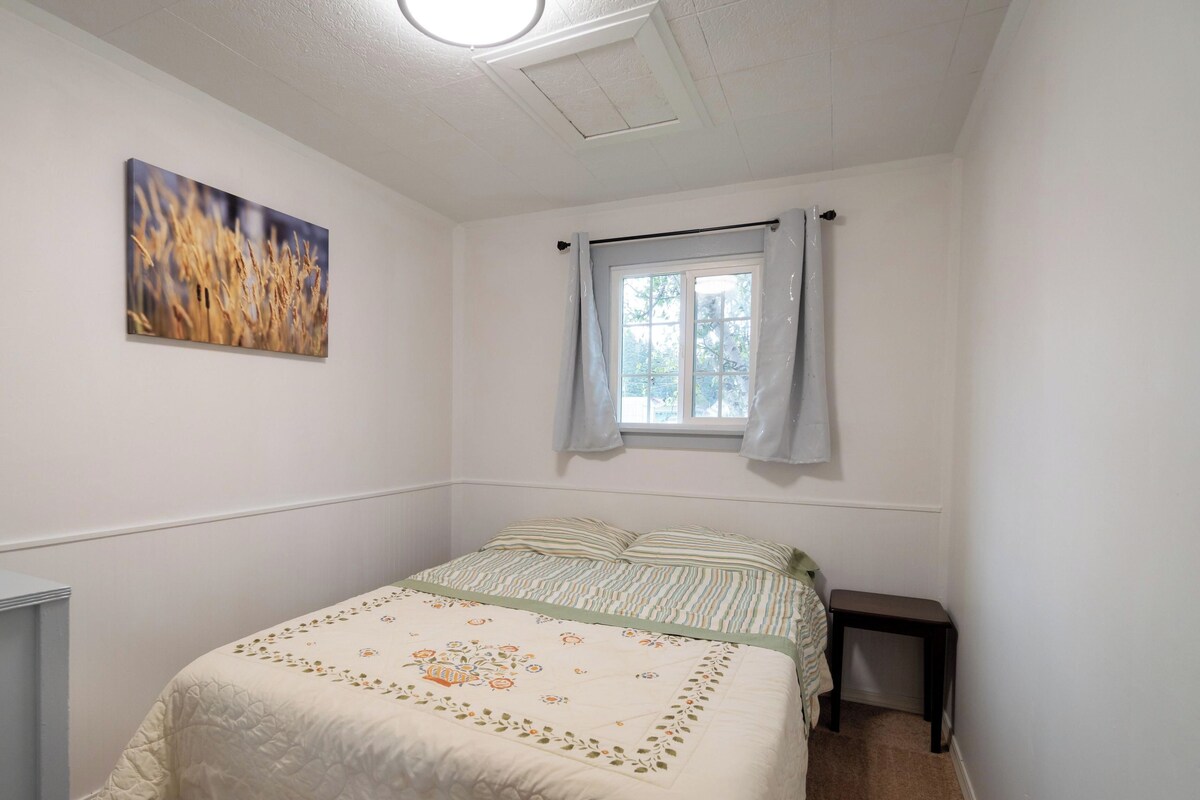 A bedroom with a bed, a window with curtains, a painting on the wall, and a ceiling light.
