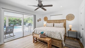Een slaapkamer met een groot bed, een plafondventilator, een bankje, een nachtkastje met een lamp, en uitzicht op een buitenzwembad.