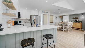 Een moderne keuken met witte kasten, een centraal eiland met krukken, een eethoek met tafel en stoelen, en een bordje met de tekst 'BEACH HOUSE'.