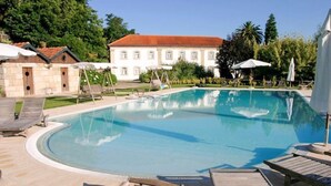Une piscine entourée de chaises longues et de parasols, elle-même bordée d’un jardin et d’un bâtiment doté d’un toit en tuiles.
