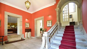 Un magnifique escalier recouvert d’un tapis rouge, orné de balustrades en pierre et surmonté d’une grande fenêtre en arc de cercle.