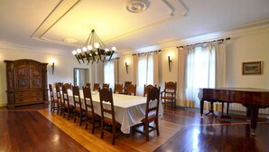 Une salle à manger avec une longue table, des chaises en bois, un lustre, un piano à queue et un buffet en bois.