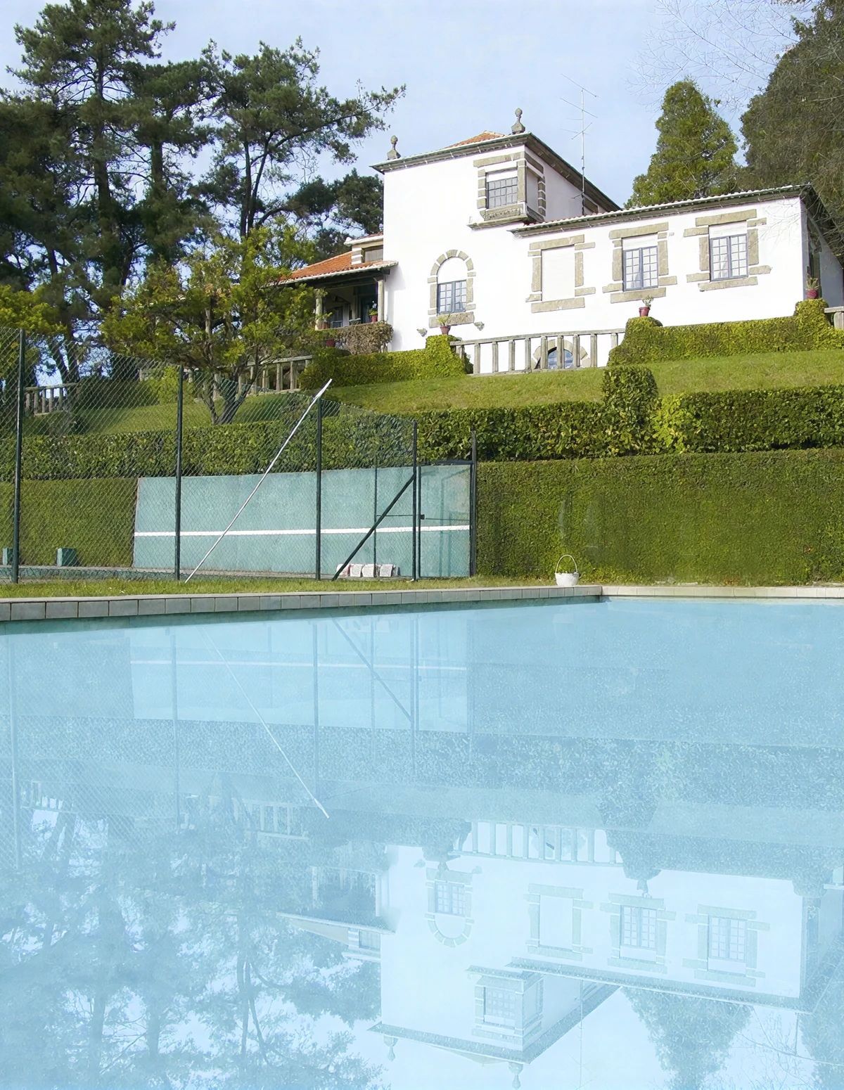 Une piscine dont l’eau est si claire qu’elle reflète parfaitement l’environnement, entourée d’une clôture et d’un beau massif de haies bien entretenu.