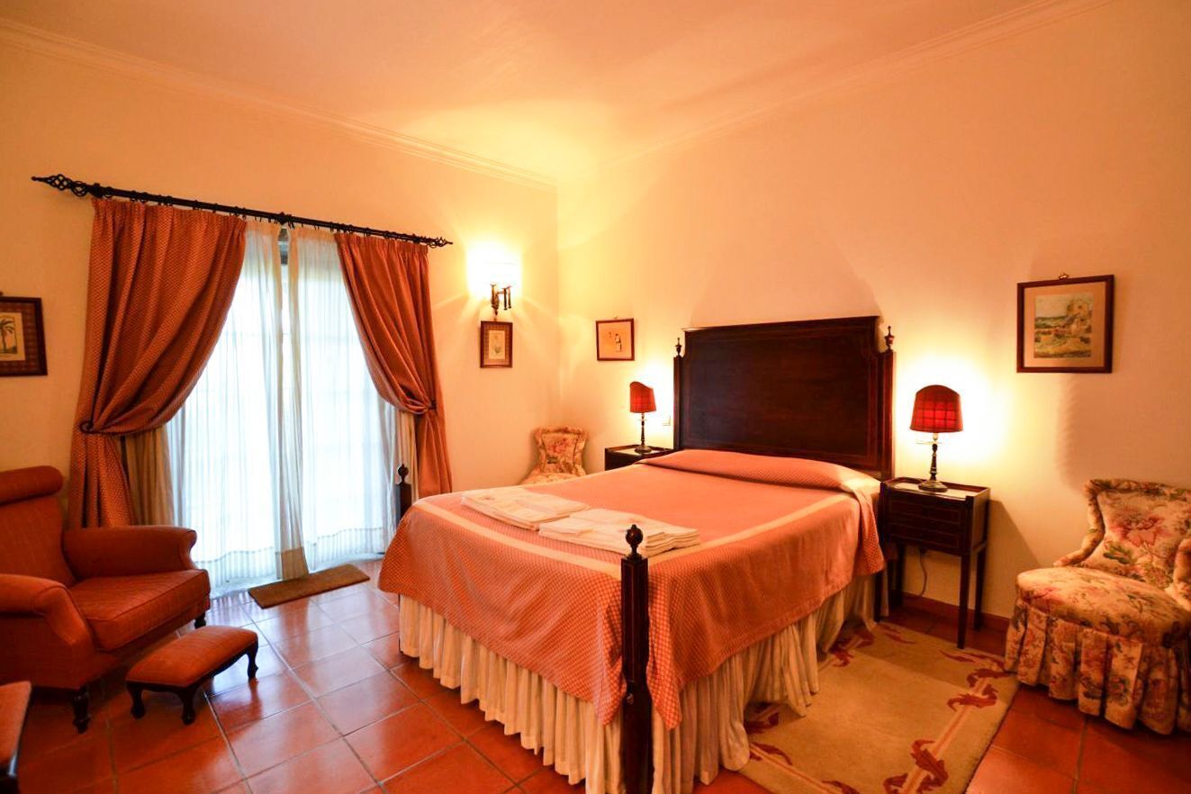 Une chambre à coucher avec un grand lit, une chaise, une table de chevet et une fenêtre avec des rideaux.