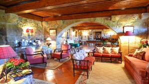 Un intérieur chaleureux avec des murs en pierre, des poutres en bois et une arche centrale qui mène à un espace repas. On y trouve plusieurs coins salon avec des canapés et des fauteuils, ainsi qu’une table agrémentée d’une lampe et de fleurs.