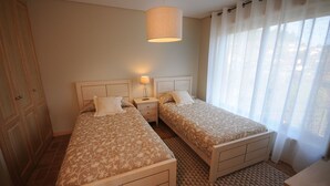A bedroom with two beds, a nightstand, a lamp, and a window with sheer curtains.