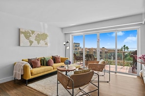 A modern living room with a yellow sofa, wicker chairs, a world map artwork, and a balcony with a view of buildings and palm trees.