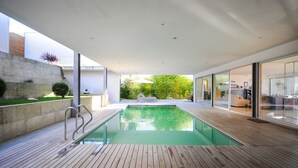 A modern indoor swimming pool with a wooden deck, glass walls, and a view of a lush garden.