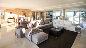 A modern living room with a large sectional sofa, a glass coffee table, and a view of the outdoors through large windows.