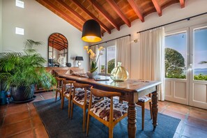 A dining area with a wooden table, chairs with patterned cushions, a hanging light fixture, and a view through a glass door. - Blanco De Caza - Three Bedroom Villa, Sleeps 6 (Alcaucín)