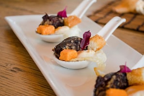 A white rectangular plate with three spoons containing food items, including what appears to be a dark-colored meat, orange cubes, and white cream, garnished with purple leaves.