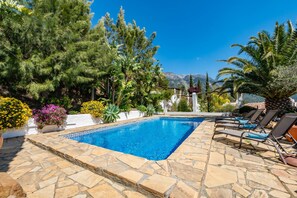 A swimming pool with stone tiles, surrounded by various plants and trees, with a mountain view in the background. - Blanco De Caza - Three Bedroom Villa, Sleeps 6 (Alcaucín)