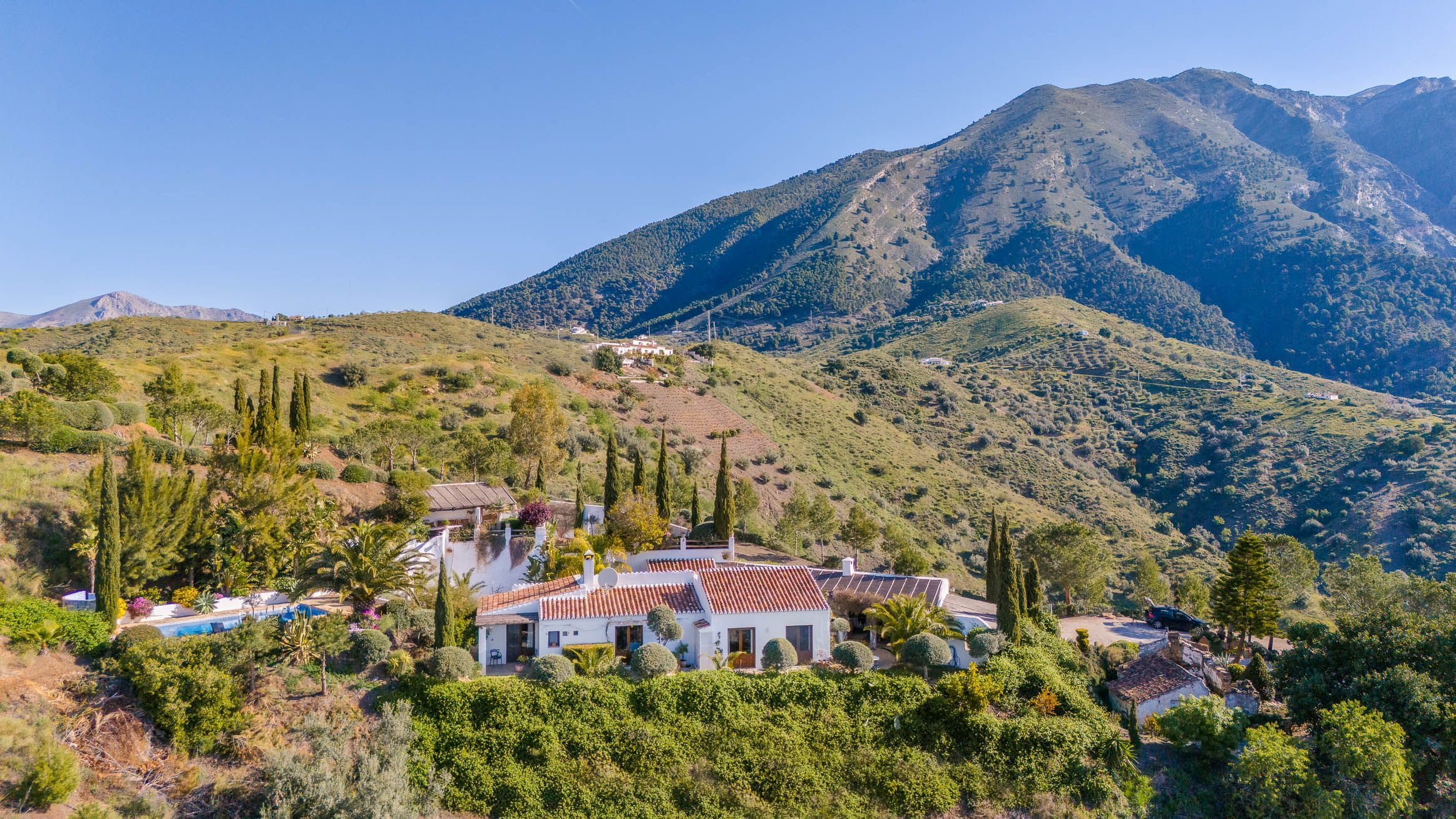 Une propriété située sur une colline, avec un bâtiment blanc doté d’un toit recouvert de tuiles rouges, entourée d’une végétation luxuriante et offrant une vue sur les montagnes en arrière-plan.