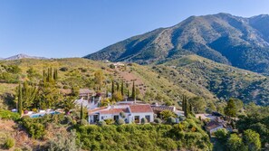 A hillside property with a white building featuring a red-tiled roof, surrounded by lush greenery and a mountain backdrop. - Blanco De Caza - Three Bedroom Villa, Sleeps 6 (Alcaucín)