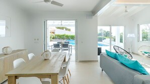 Ein modernes Wohnzimmer mit einem Holzesstisch, weißen Stühlen, einem grauen Sofa mit Türkis-Kissen und Blick auf einen Swimmingpool und Grünflächen.