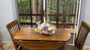 Une table à manger agrémentée d’un centre de table floral, entourée de chaises en bois, dans une pièce dotée de grandes fenêtres donnant sur une forêt.