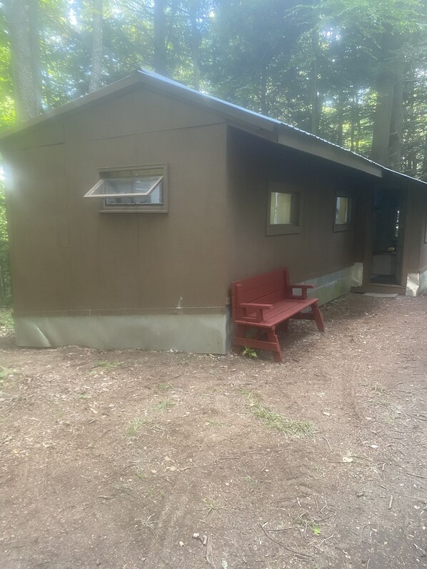 A small, single-story building with a sloped roof, a bench outside, and a window on the side.