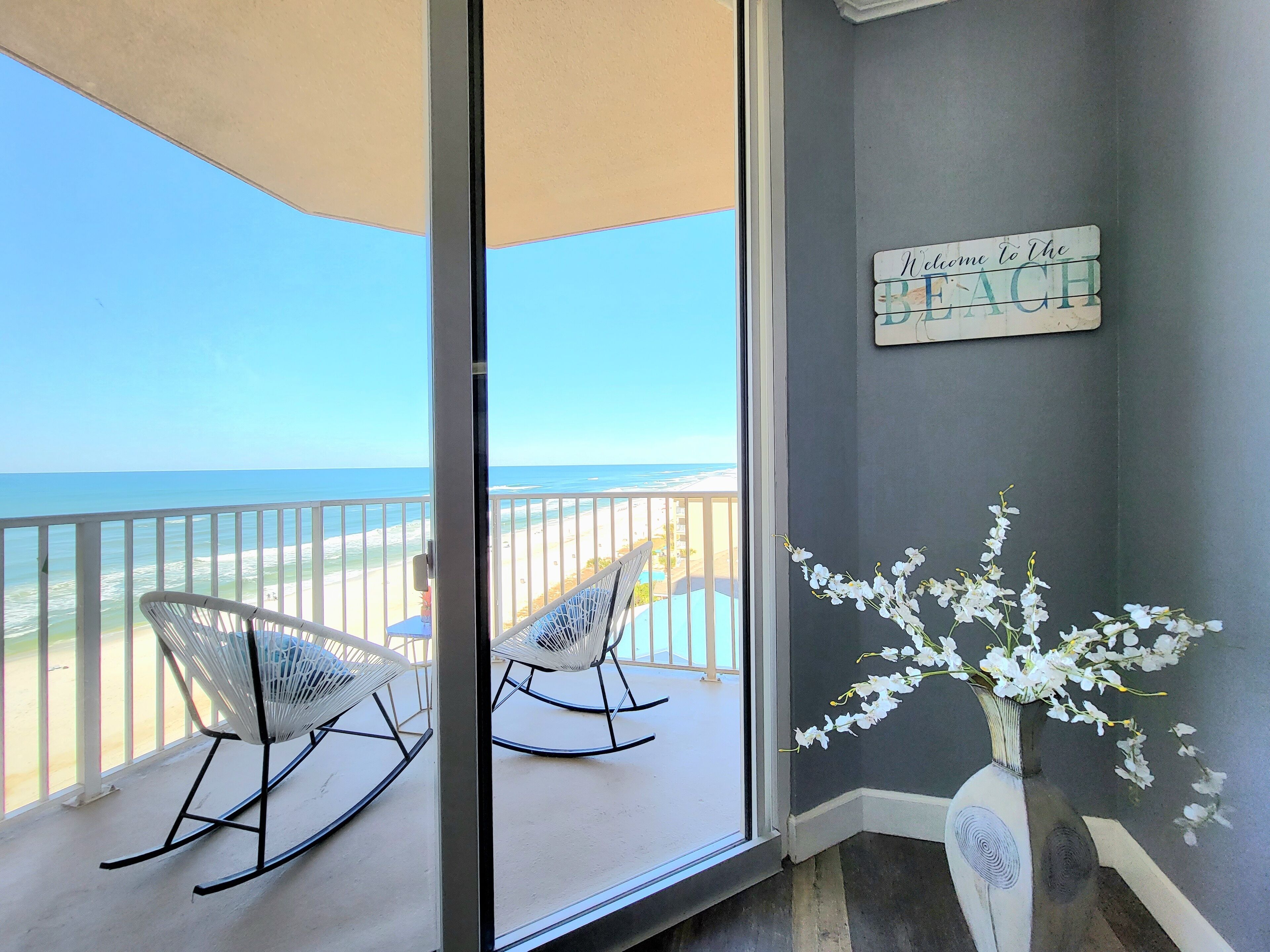 A balcony with a view of the beach, two rocking chairs, a vase with white flowers, and a sign that reads "Welcome to the BEACH".