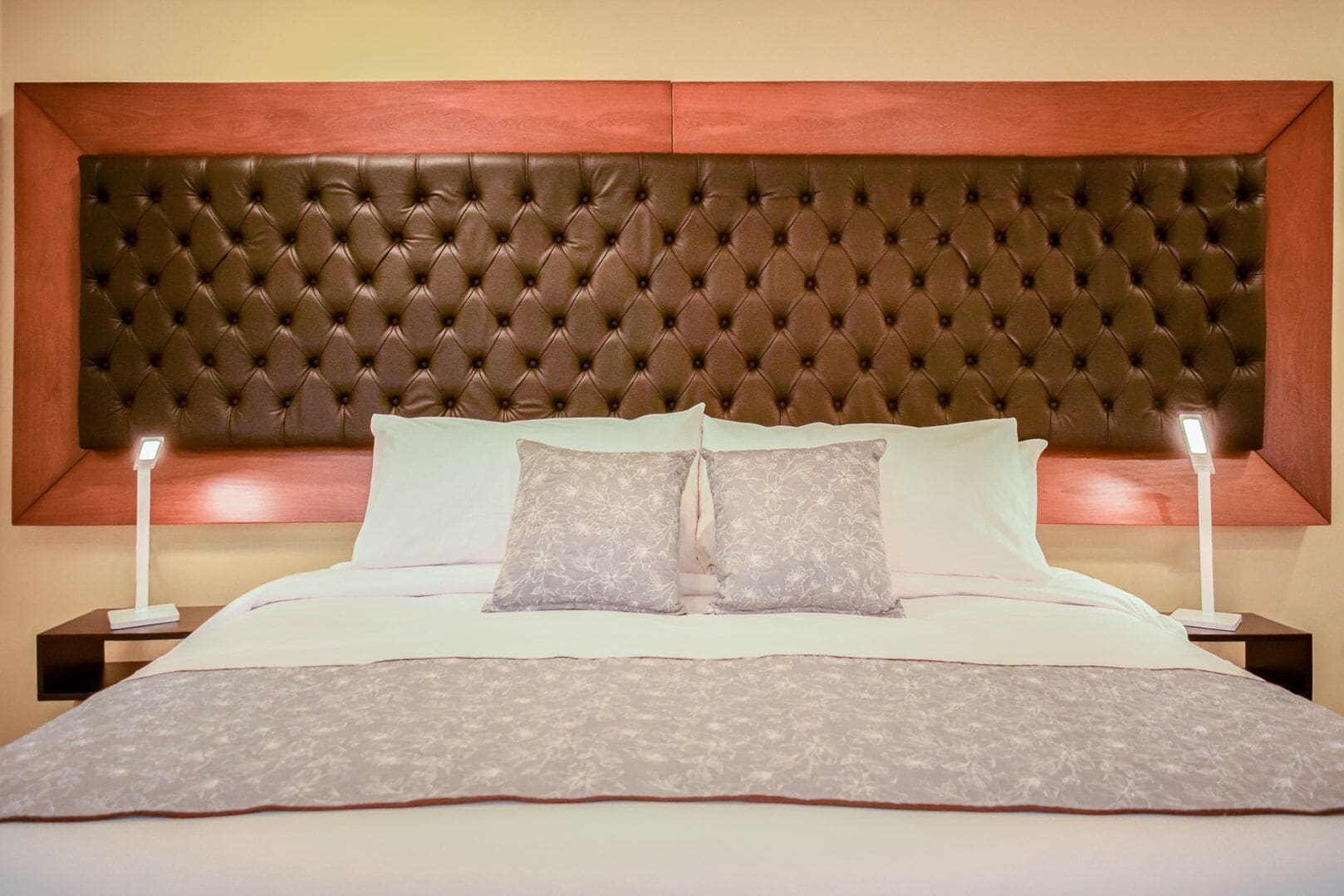 Ein Hotelzimmer mit einer gepolsterten Kopfstütze, einem großen Bett mit weißen Laken, zwei Nachttischen mit Lampen und floralen Kissen.