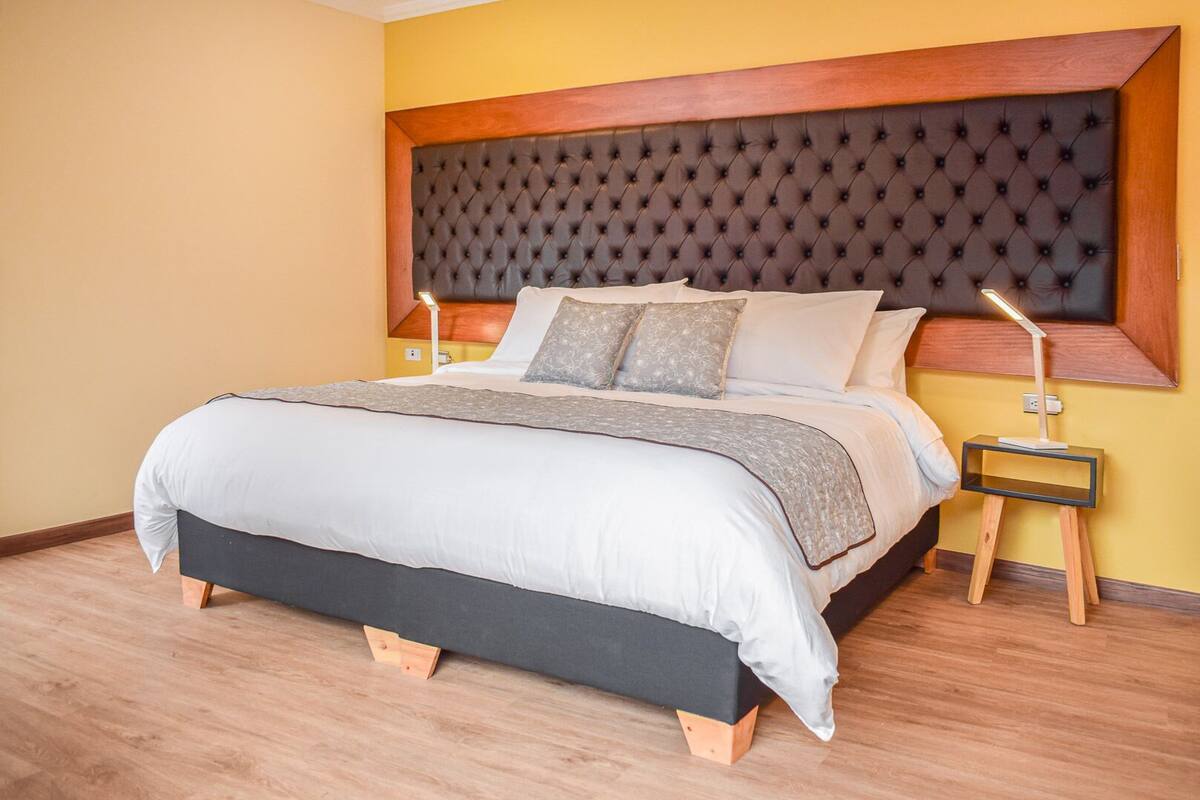 Una cama bien hecha con cabecera capitonada, una mesita de noche con lámpara y suelo de madera.