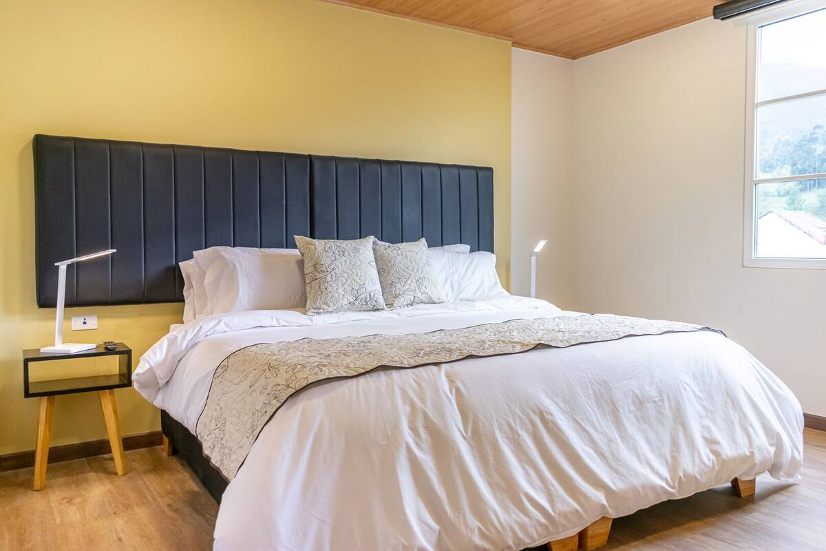 Una cama bien hecha con cabecera oscura, ropa de cama blanca y una mesita de noche con lámpara.