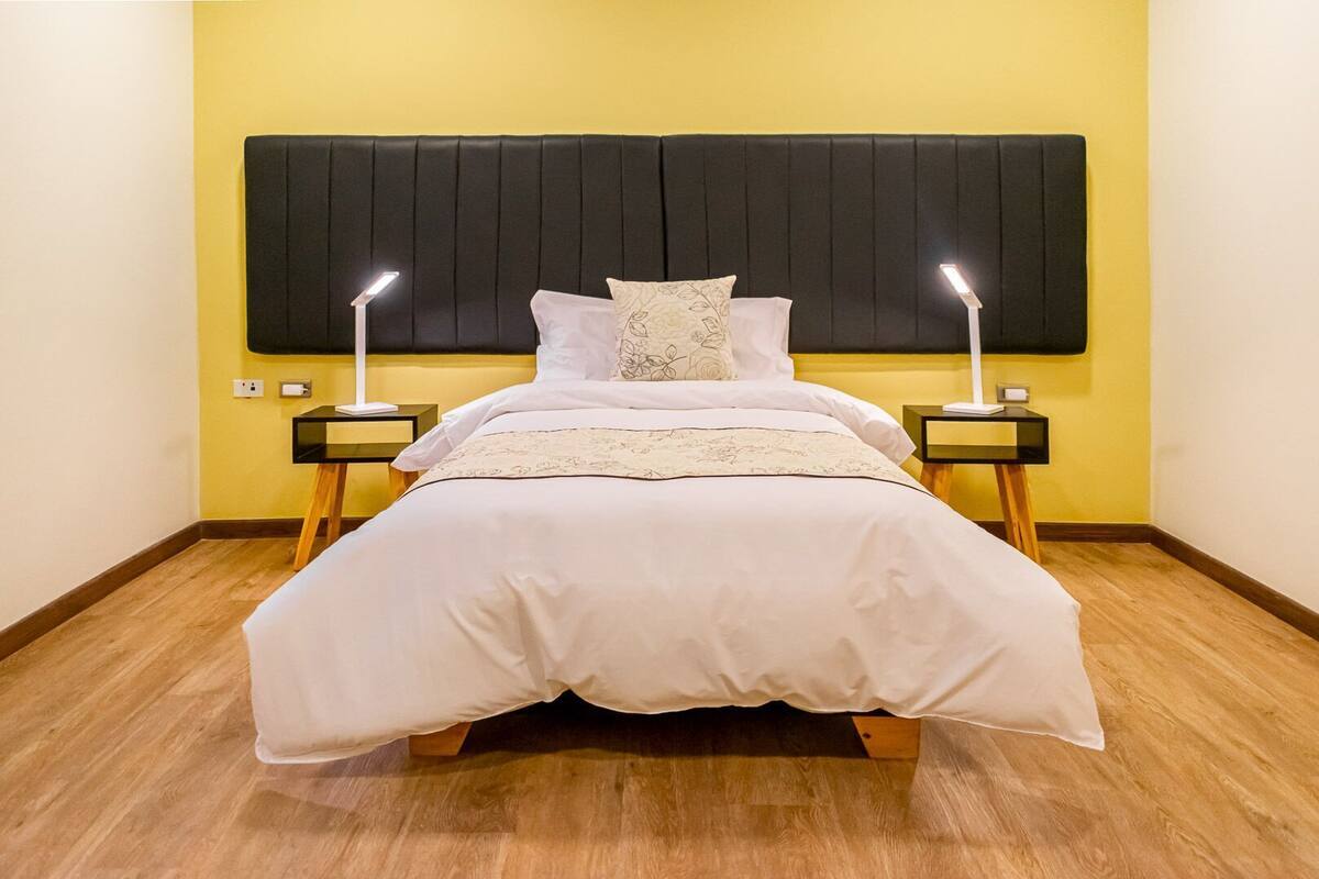 Una cama con cabecera negra, dos lámparas de noche y una pared amarilla.