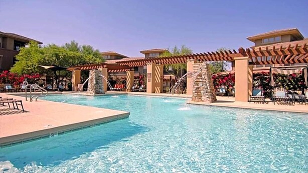 A swimming pool with a clear blue water, surrounded by lounge chairs and umbrellas, and a stone structure in the center.