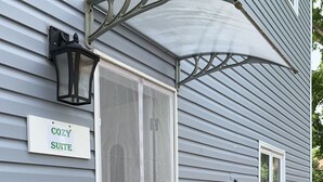 A house with a 'COZY SUITE' sign, a covered entryway, and a flower bed with pink roses. - Ambiance by the Falls Cozy Suite (Niagara Falls)