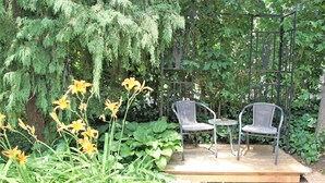 A wooden deck with two wicker chairs and a small round table, surrounded by lush greenery and flowering plants. - Ambiance by the Falls Cozy Suite (Niagara Falls)