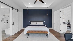 A modern bedroom with a large bed, a wooden bench, and a ceiling fan. - Lakefront 5BR w/ Pool, Hot Tub & Game Room. Sleeps 24. Family-Friendly (Seguin)