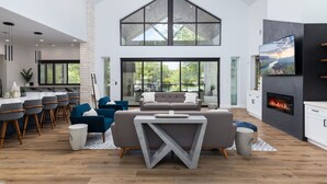 A spacious living area with a high ceiling, a fireplace, a large TV, and a modern kitchen with bar seating. - Lakefront 5BR w/ Pool, Hot Tub & Game Room. Sleeps 24. Family-Friendly (Seguin)
