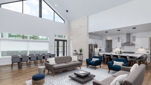 A spacious living area with a high ceiling, a dining area with a long table and chairs, a kitchen with modern appliances, and a large window providing natural light. - Lakefront 5BR w/ Pool, Hot Tub & Game Room. Sleeps 24. Family-Friendly (Seguin)