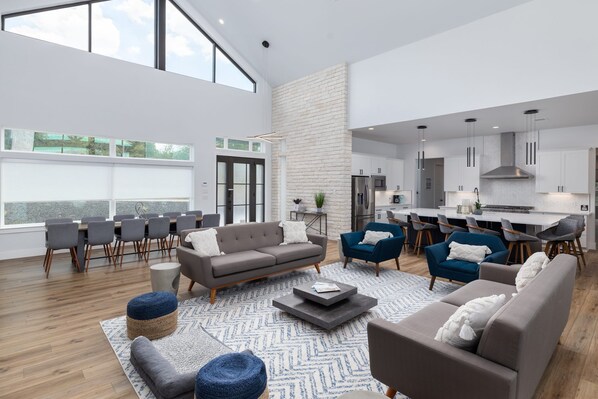 Een ruime woonkamer met een hoge plafond, een eethoek met een lange tafel en stoelen, een keuken met moderne apparatuur, en een groot raam dat voor natuurlijk licht zorgt.