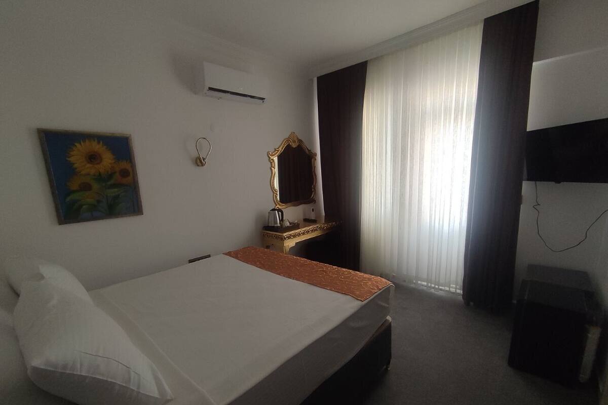 Ein Hotelzimmer mit einem Bett, einem Nachttisch, einem Spiegel, einem Sonnenblumengemälde und einem Fenster mit Vorhängen.