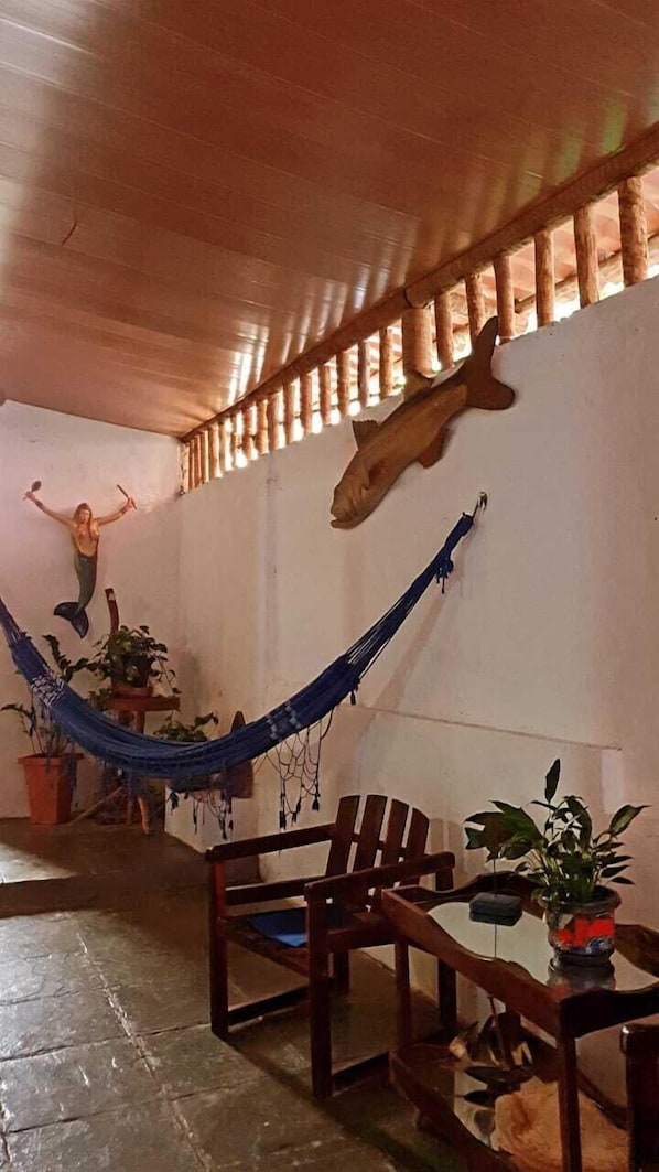 A room with a hammock, wooden chairs, a table with a plant, and a mermaid decoration on the wall. - Pousada Passarte Biquinha (Cairu)